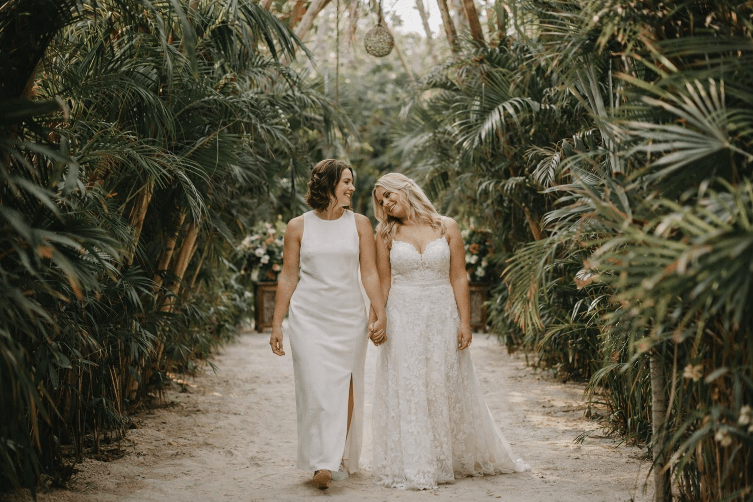 Two brides walking hand in hand through a jungle ceremony path at a Riviera Maya destination wedding